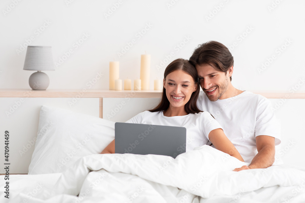 Happy european millennial man and woman hugging, sitting on bed, use laptop in white bedroom interior Happy european millennial man and woman hugging, sitting on bed, use laptop in white bedroom interior