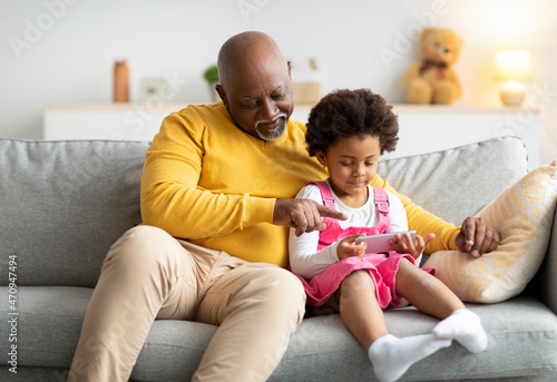 Smiling busy african american senior grandfather and little girl sitting on couch, granddaughter playing in online game photo