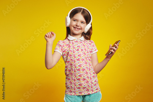 Cheerful young girl with white bluetooth headphones, listening music from smartphone and dancing, standing isolated over yellow background