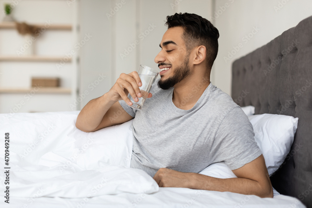 Smiling Arab Guy Drinking Mineral Water From Glass While Sitting In Bed Smiling Arab Guy Drinking Mineral Water From Glass While Sitting In Bed