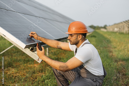 Competent technician in safety helmet and glasses measuring amperage of solar panels with multimeter. Indian man repairing photovoltaic cells outdoors. Green energy concept. photo