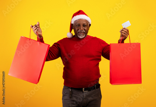 Happy senior black man in Santa Claus hat holding credit card and Christmas gift bags with mockup for your brand logo