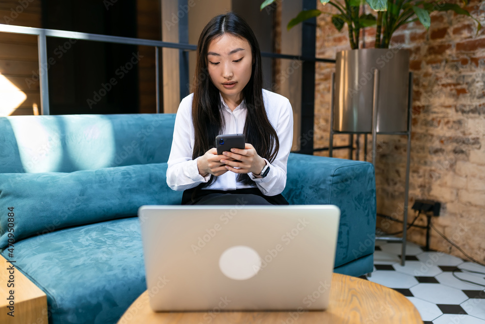 Naklejka premium Asian woman doing online shopping using the phone while sitting at the table in front of the laptop