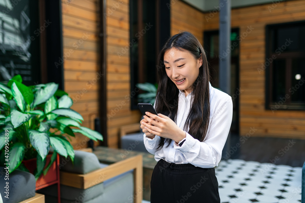 Asian smiling young woman stands in the corridor and looks into a mobile phone decides matters.