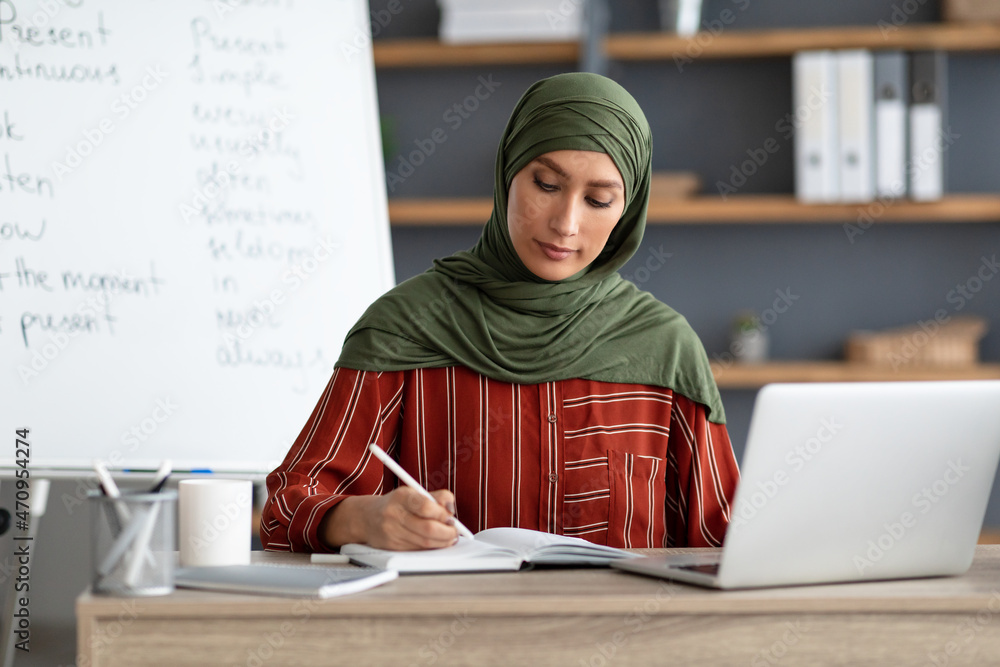 Muslim teacher in headscarf sitting at desk, writing, using pc Muslim teacher in headscarf sitting at desk, writing, using pc