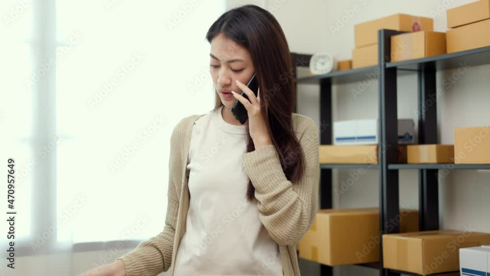 Busy Young asian business woman using mobile phone call receiving purchase order and check product on stock near the package shelf on background. Ecommerce business and shopping online concept. Busy Young asian business woman using mobile phone call receiving purchase order and check product on stock near the package shelf on background. Ecommerce business and shopping online concept.