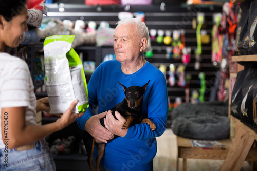 Old man choosing dog food for his miniature pinscher in pet shop. Saleswoman helping him with it.