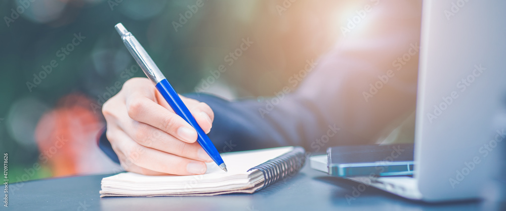 Woman hand writing on a notepad with a pen and she is holding a cell phone in the office.