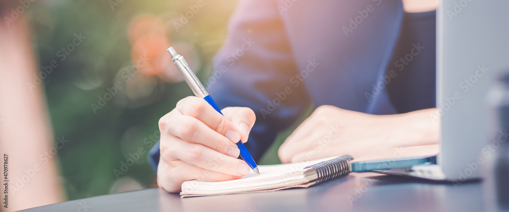 Woman hand writing on a notepad with a pen and she is holding a cell phone in the office.
