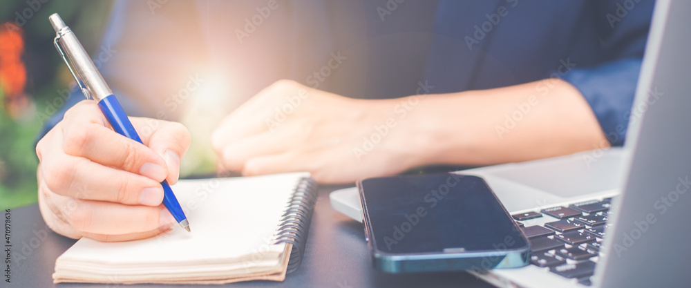 Woman hand writing on a notepad with a pen and she is holding a cell phone in the office.