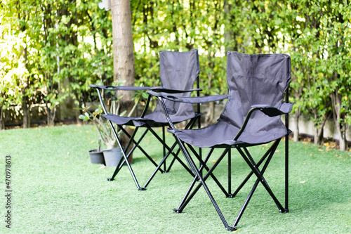 An oblique view, selective focus of two black empty canvas folding camping chairs placed on a green grass carpet near a tree and plant pots surrounding by blurred small trees lining as a lush wall.