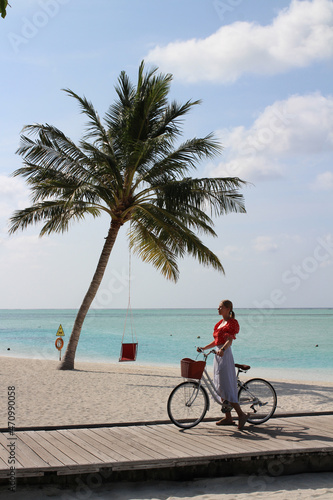 A girl in a red blouse and a white skirt stands with a bicycle on a wooden pier against the background of a palm tree with a hammock  a white sand beach and the ocean in the Maldives.