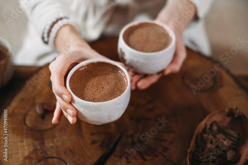 Hot handmade ceremonial cacao in white cups. Woman hands giving craft cocoa, top view on wooden table. Organic healthy chocolate drink prepared from beans, foam Giving cup on ceremony cozy atmosphere photo