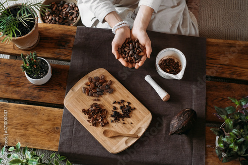 Woman hands holding organic cacao beans on wooden table, cocoa nibs, artisanal chocolate making in rustic boho style for ceremony. Degustation, Chocolate making with pounder close-up top view
