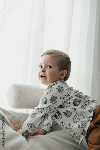 A happy little child in a white jacket and brown pants lies comfortably on a white soft sofa and laughs against the background of a large bright window. 