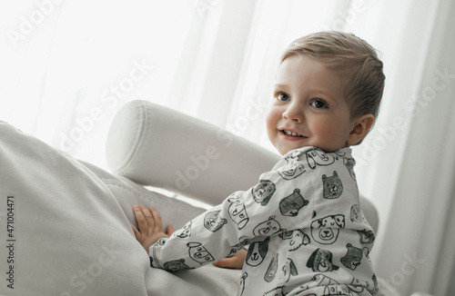 A happy little child in a white jacket and brown pants lies comfortably on a white soft sofa and laughs against the background of a large bright window. 
