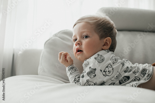 A happy little child in a white jacket and brown pants lies comfortably on a white soft sofa and laughs against the background of a large bright window. 