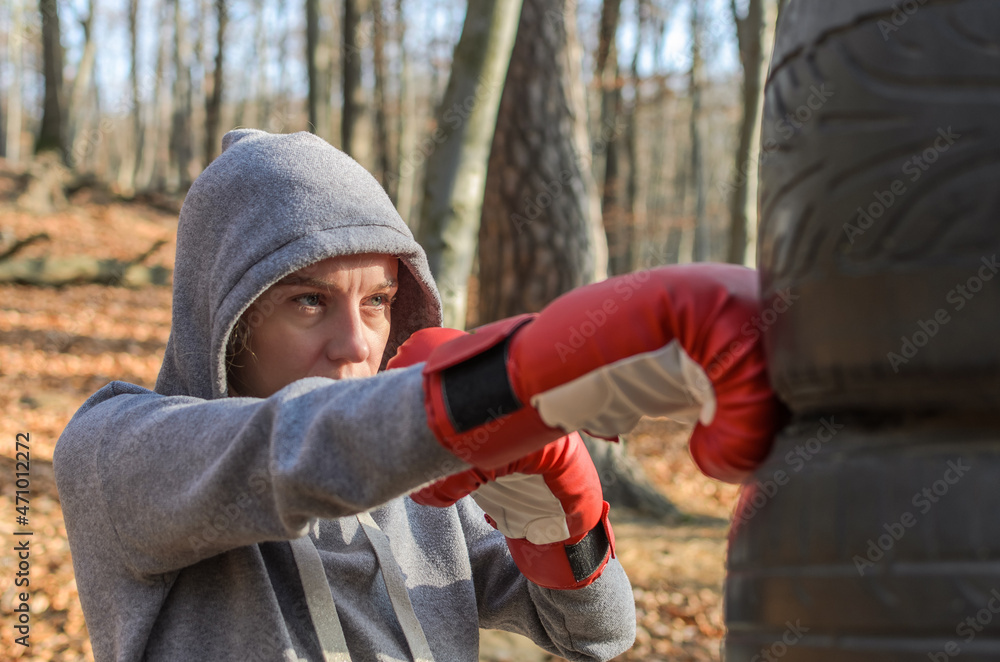 Young charming woman in red boxing gloves, dressed in a tracksuit with a  hood, in a boxing training in the forest outdoors, beats a homemade  punching bag from car tires Stock Photo