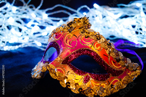 Purple and yellow carnival mask on black silk. There are festive white lights in the background.