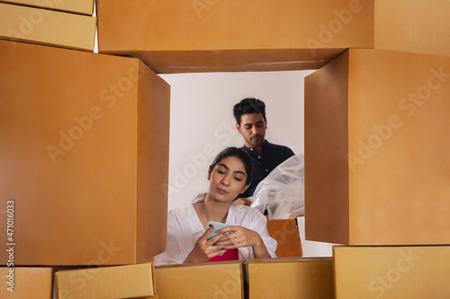 Girl watching Smartphone and boy standing with box behind stacking of cardboard boxes