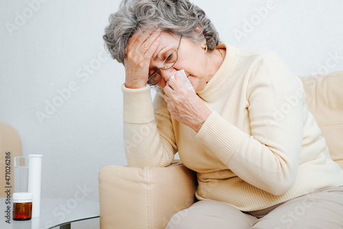 Senior sick woman with flu, runny nose and headache sitting by atable with pills alone at home. Lonely elderly woman and virus, allergy, disease concept. Healthcare, drug treatment photo