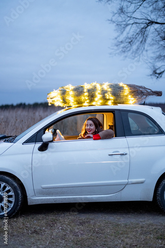 Fototapeta Naklejka Na Ścianę i Meble -  Woman driving car with a illuminated Christmas tree on a rooftop. Concept of New Year preparation. Idea of fairy tale and festive mood in wintertime. Side view, blurred photo technic
