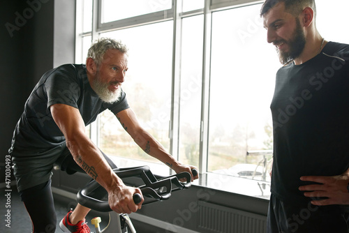 Elderly man on bike with young trainer in gym