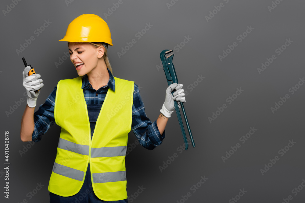 Angry builder in helmet screaming in walkie talkie and holding pipe wrench isolated on grey