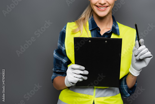Cropped view of cheerful blonde builder in gloves and safety vest holding pen and clipboard isolated on grey © LIGHTFIELD STUDIOS