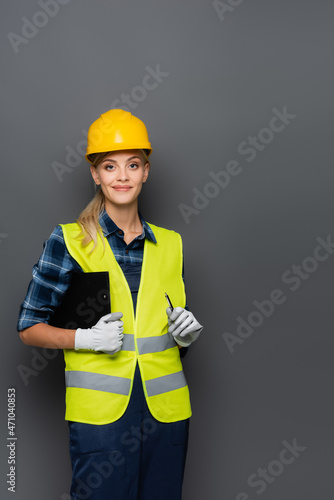 Cheerful builder in hardhat looking at camera while holding clipboard and pen isolated on grey