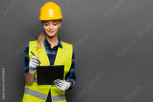 Smiling builder in hardhat and safety vest holding pen and clipboard isolated on grey