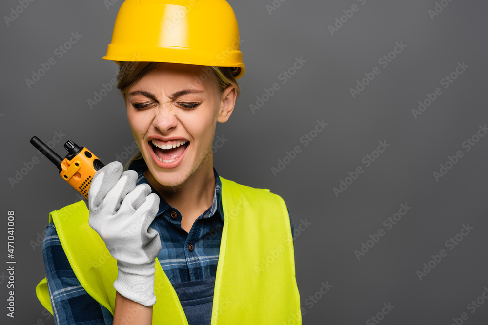 Angry builder in hardhat screaming while holding walkie talkie isolated on grey Angry builder in hardhat screaming while holding walkie talkie isolated on grey