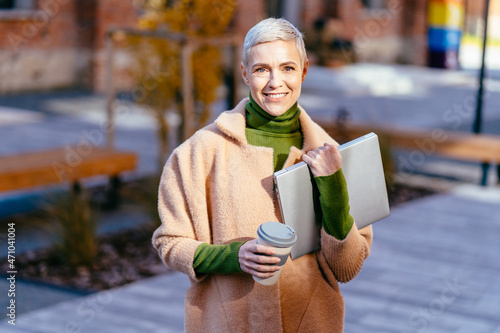 Elegant blond short hair 40-45 years old woman against brick wall outdoor with silver laptop and takeaway reusable coffee cup in hands. Remote working concept with flexible lifestyle. photo