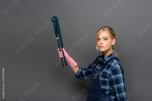 Blonde plumber holding pipe wrench and looking at camera on grey background
