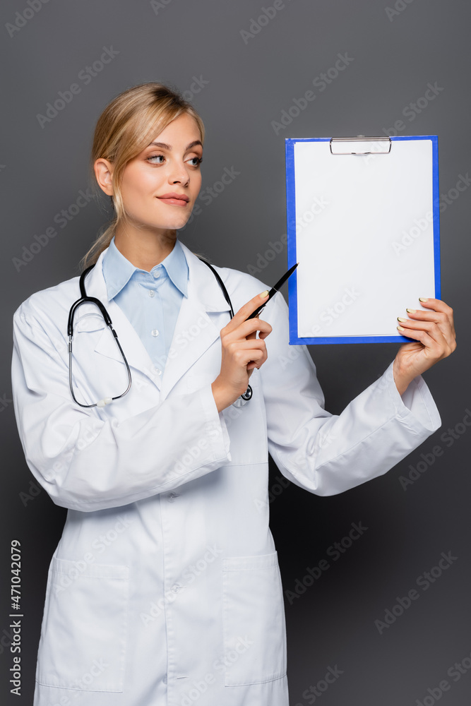 Young doctor pointing at clipboard with copy space isolated on grey Young doctor pointing at clipboard with copy space isolated on grey