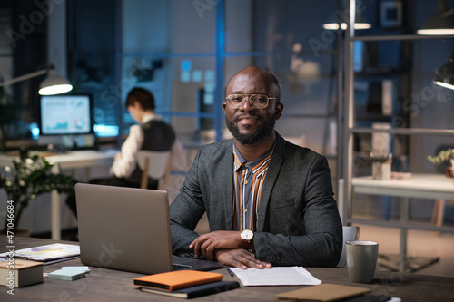 Portrait of African businessman in eyeglasses smiling at camera while working at his workplace with laptop till late evening at office