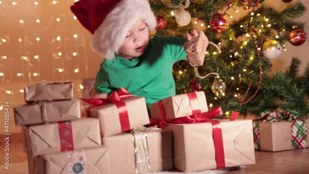 caucasian 3 year child kid boy in green t-shirt and red Santa hat sitting on floor under decorated Christmas tree putting wrapped presents gift boxes in big pile stack. waiting preparing, celebrating caucasian 3 year child kid boy in green t-shirt and red Santa hat sitting on floor under decorated Christmas tree putting wrapped presents gift boxes in big pile stack. waiting preparing, celebrating