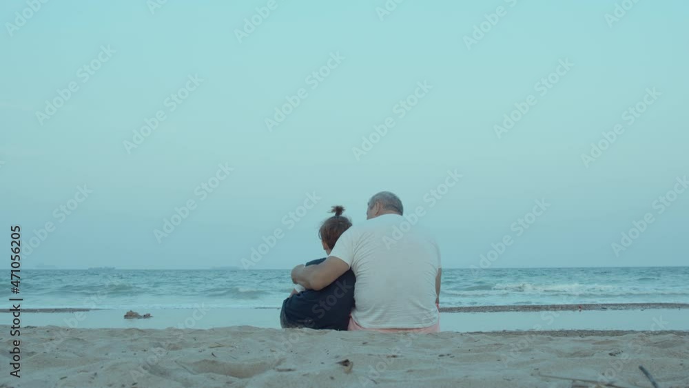 Wide shot - grandfather hugs his grandson, tells him something, sitting on the sandy shore, near the sea, looking at the waves. Shot from the back Wide shot - grandfather hugs his grandson, tells him something, sitting on the sandy shore, near the sea, looking at the waves. Shot from the back