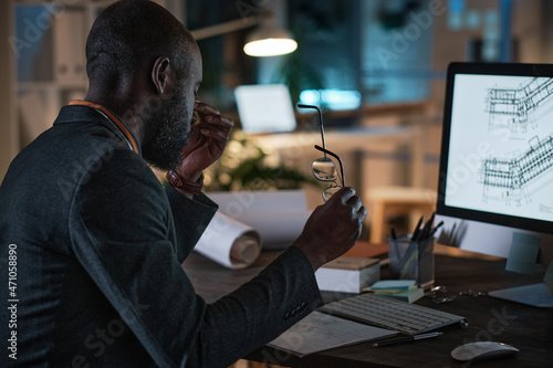 Tired African architect sitting at the table in front of the computer monitor with blueprint while working at office till night