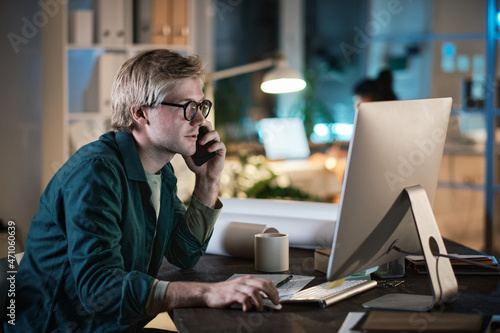 Young businessman in eyeglasses working on computer and talking on mobile phone while sitting at the table and working late at office