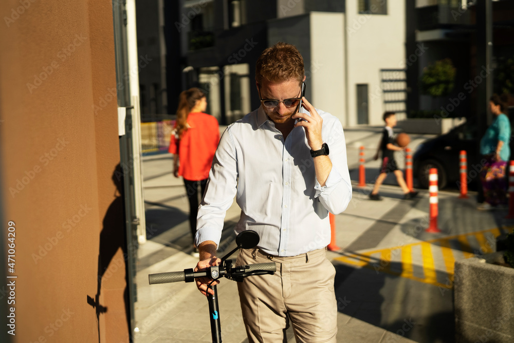 Young happy businessman with electric scooter outdoors. Portrait of handsome man talking to the phone.