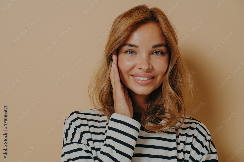 Portrait of good looking young woman touches face gently smiles toothily looks directly at camera Portrait of good looking young woman touches face gently smiles toothily looks directly at camera