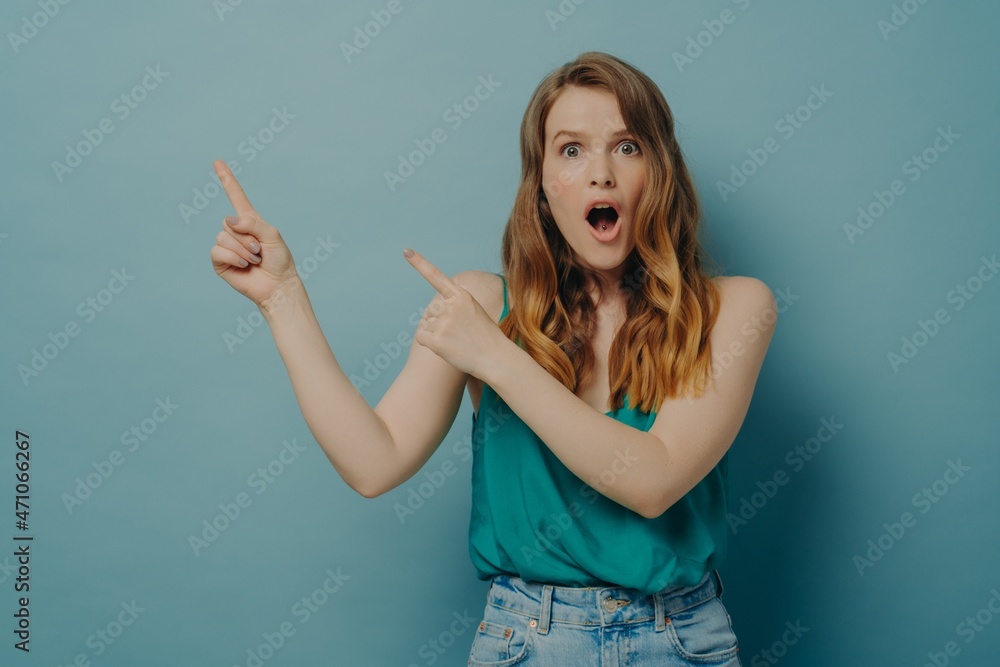 Surprised young woman in casual clothes pointing to right side with index fingers, isolated on blue Surprised young woman in casual clothes pointing to right side with index fingers, isolated on blue