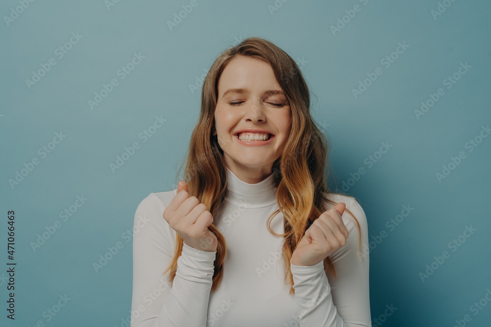 Excited young lady clenched fists waiting for miracle with eyes closed, isolated on blue background Excited young lady clenched fists waiting for miracle with eyes closed, isolated on blue background