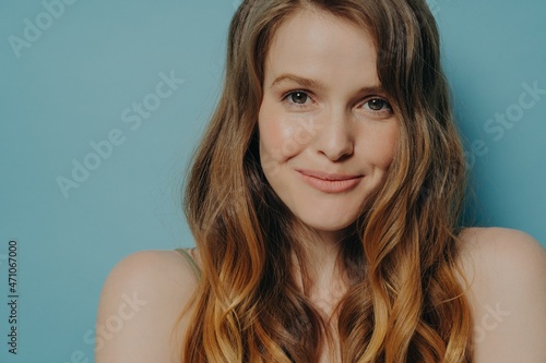 Studio shot of charming shy young woman with wavy brown hair looking at camera with slight smile