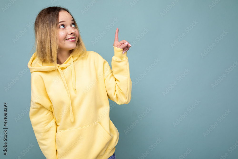 Portrait of young beautiful smiling hipster blonde woman in trendy yellow hoodie. Sexy carefree female person posing isolated near blue wall in studio. Positive model with natural makeup. Copy space Portrait of young beautiful smiling hipster blonde woman in trendy yellow hoodie. Sexy carefree female person posing isolated near blue wall in studio. Positive model with natural makeup. Copy space
