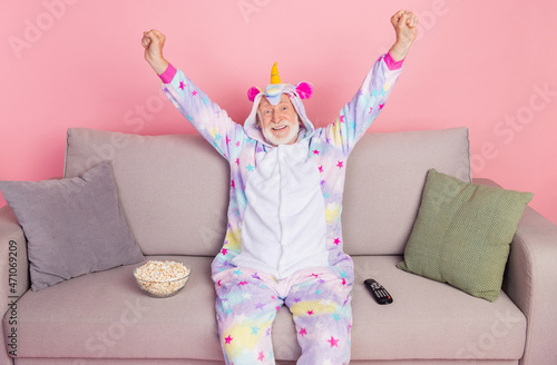 Photo of funny lucky retired man nightwear overall sitting couch watching movie eating pop corn rising fists isolated pink color background © deagreez