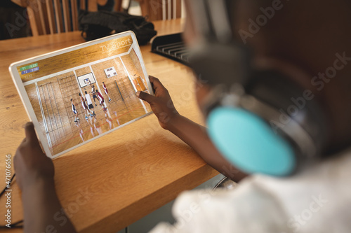 African american boy wearing headphones at home watching basketball game on tablet