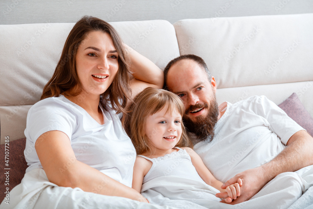 Young family play spend time together hugs on couch in morning. Mother Father and Daughter child in white t-shirts pajamas have fun in Morning Routine at home in bedroom. 