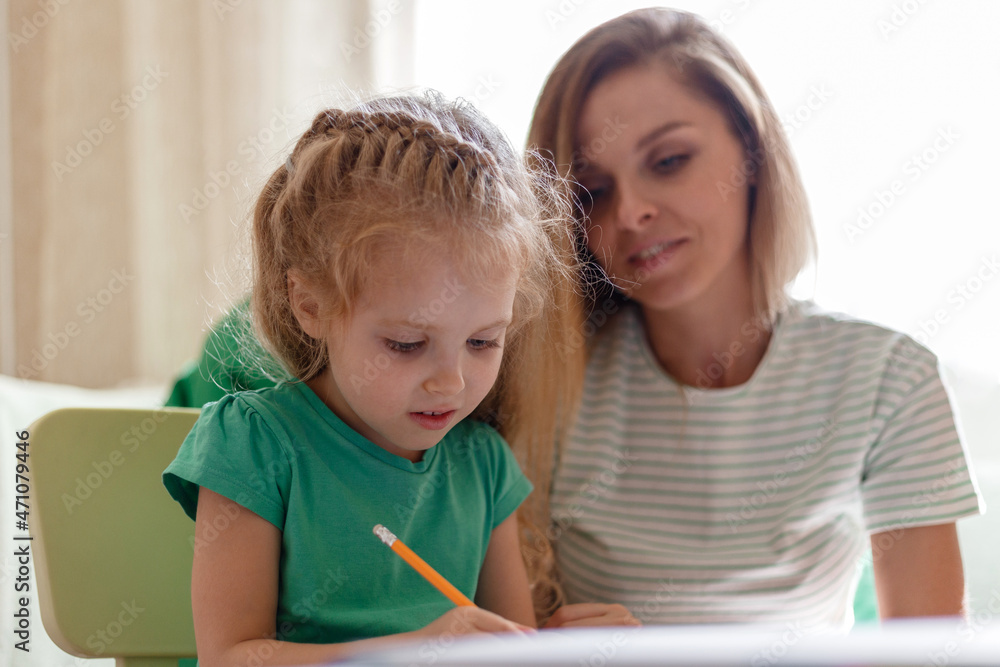 Mother helps preschooler girl daughter to do homework. Caucasian blonde smart child kid 5 years do homework Learn to write with mom 30s together. Homeschooling and Kindergarten Concept Mother helps preschooler girl daughter to do homework. Caucasian blonde smart child kid 5 years do homework Learn to write with mom 30s together. Homeschooling and Kindergarten Concept